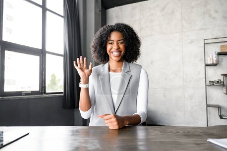 Photo for With a radiant smile and friendly wave, African-American woman greets her colleagues during a video call. Modern office provides an ideal backdrop for virtual business meetings and collaborative work - Royalty Free Image