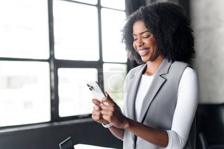Une femme afro-américaine joyeuse dans un élégant blazer gris regarde son smartphone avec un sourire éclatant dans un cadre de bureau moderne, respirant le professionnalisme et l'accessibilité.