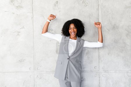 An exuberant African-American businesswoman celebrates with her arms raised against a textured gray background, embodying success and joyful achievement in the business world.