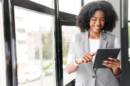 With a confident stance and a tablet in hand, the businesswoman looks down at her device with an engaging smile, surrounded by the modern comforts of a chic office space.