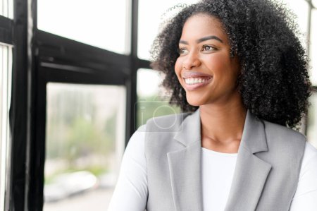 A beaming African-American businesswoman looks out the window with an optimistic gaze, reflecting a forward-thinking attitude in a contemporary, well-lit office setting.