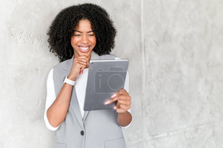Photo for A young African-American businesswoman with an infectious grin uses a tablet, showcasing a balance of friendliness and professionalism against a minimalistic concrete backdrop - Royalty Free Image