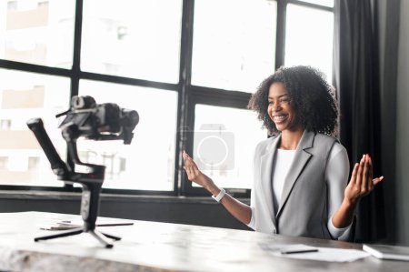 With a warm smile and open hands, businesswoman conveys a sense of approachability and friendliness, inviting interaction in an educational vlog or webinar from her office, recording vlog or classes