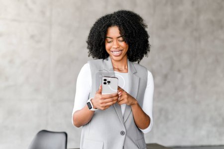 Photo for An engaging African-American businesswoman interacts with her smartphone, her expression reflecting the ease and efficiency of modern business communications within a contemporary office setting. - Royalty Free Image
