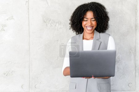 Eine fröhliche afroamerikanische Geschäftsfrau hält einen Laptop in der Hand, ihr ansteckendes Lächeln suggeriert Produktivität und Leichtigkeit im Umgang mit Technologie in einem Unternehmensumfeld..