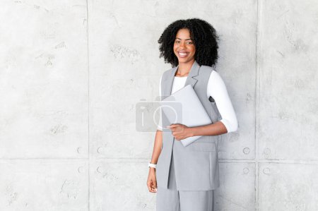 Photo for Exuding professionalism, an African-American woman stands confidently with a laptop in hand, wearing a business-casual outfit that perfectly blends modern work attire with comfort - Royalty Free Image