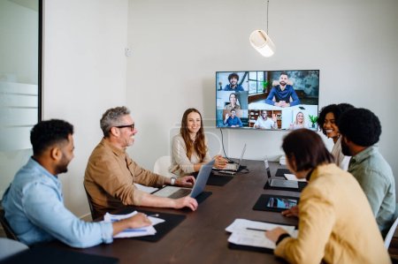 Se captura una animada y productiva reunión de equipo donde los colegas se conectan a través de una pantalla grande, mostrando una mezcla de colaboración en persona y remota. Concepto de reunión virtual