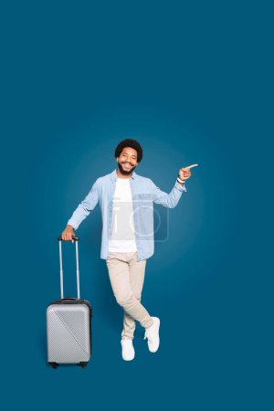 The spirited young traveler points happily to the side, standing with a suitcase and an expression of joy, suggesting recommendations or travel options, isolated on blue