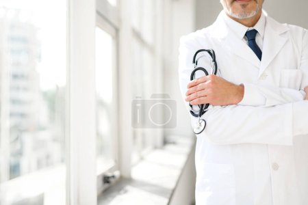 Photo for Cropped photo with focus at a stethoscope in hands of senior doctor. Poised MD in a white coat stands with crossed arms embodying confidence and experience against a backdrop of a bright clinic window - Royalty Free Image