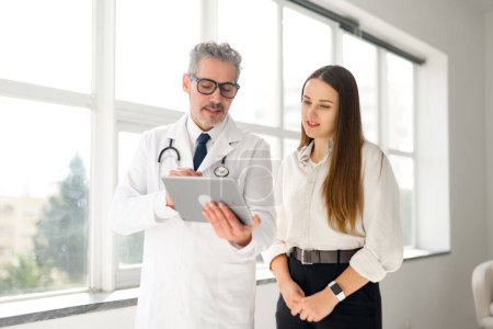 En esta imagen, un médico maduro con una tableta digital discute opciones de atención médica con una paciente joven en una habitación bien iluminada, ilustrando la dinámica de la consulta médica moderna
