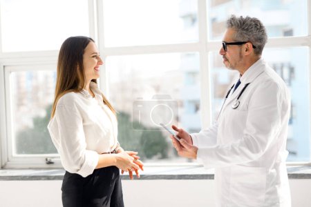 Téléchargez les photos : Un médecin chevronné aux cheveux gris engageant une conversation amicale avec une jeune patiente près d'une fenêtre, reflétant un cadre de soins de santé à la fois personnel et imprégné de lumière du jour, vue de côté - en image libre de droit
