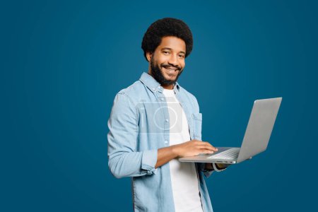 Friendly young man using laptop, capturing the blend of modern worker of professionalism and approachability in a digital environment. Brazilian male freelancer or student with laptop isolated on blue