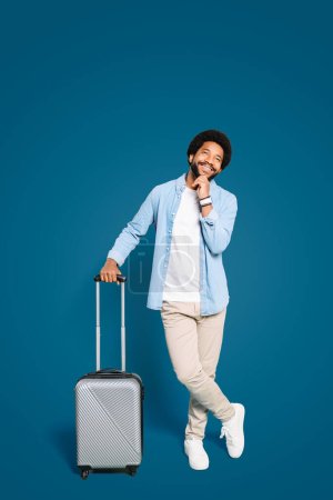 A young Brazilian man standing with suitcase in a thoughtful pose, touching his chin, the man contemplates next destination, against a blue background. Concept of travel planning and adventures