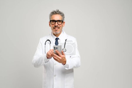 Confident senior doctor looking at the camera while using a smartphone, looking through medical apps or test results. A modern healthcare approach, integrating technology