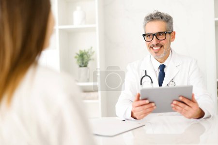 A cheerful senior doctor with grey hair and stylish glasses smiles warmly, holding a digital tablet, suggesting a modern approach to patient care. The efficiency and technological integration concept