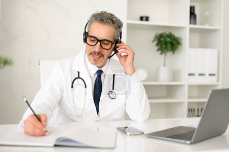 A senior grey-haired doctor in a white coat and a headset is conversing with a patient, exemplifying the personalized touch in telehealth services, attentively taking notes during virtual appointment