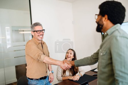 Téléchargez les photos : Un homme en chemise beige serre la main d'un collègue, signifiant un lieu de travail convivial et collaboratif, une femme en arrière-plan sourit, observant l'interaction chaleureuse - en image libre de droit