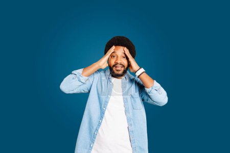 The man poses with hands on head, playful expression of surprise or shock on his face, against blue background. This image captures a spontaneous, emotive moment with a sense of humor and personality.
