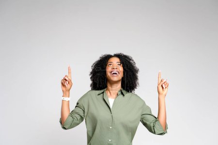 Téléchargez les photos : Une femme afro-américaine joyeuse pointe vers le haut des deux mains, regardant ravie de sa découverte, capturant un sentiment de réussite ou pointant une idée. Concept représente des moments d'inspiration - en image libre de droit