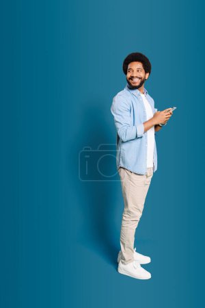 Ein fröhlicher brasilianischer Mann in Freizeitkleidung mit Smartphone steht in voller Länge isoliert auf blauem Grund und blickt auf den Kopierraum, der einen Platz für Werbung, männliche Freiberufler oder Studenten mit Telefon vorschlägt.