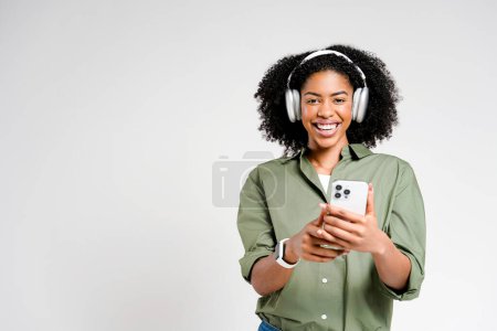 Téléchargez les photos : Une femme afro-américaine de contenu avec casque utilise son smartphone, peut-être en sélectionnant de la musique ou en assistant à une réunion virtuelle - en image libre de droit