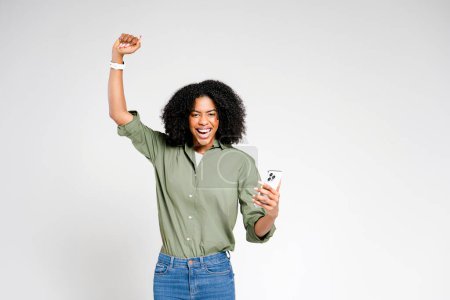 An exuberant African-American woman raises her fist in a victorious gesture while holding a smartphone in the other hand, her open-mouthed smile suggesting triumph or a significant achievement.