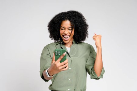 Foto de Una mujer afroamericana extática celebra mientras mira su teléfono inteligente, su puño bombeado en el aire señalando un momento de éxito o buenas noticias - Imagen libre de derechos