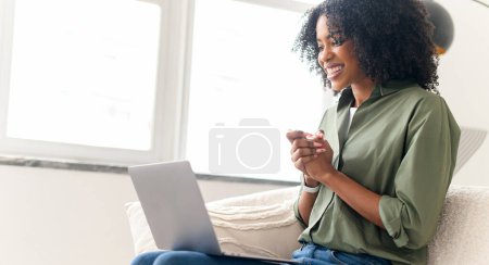 With a gleeful expression, an African-American woman gestures during a lively conversation on a video call, enjoying the comforts of her home, using laptop for virtual meeting