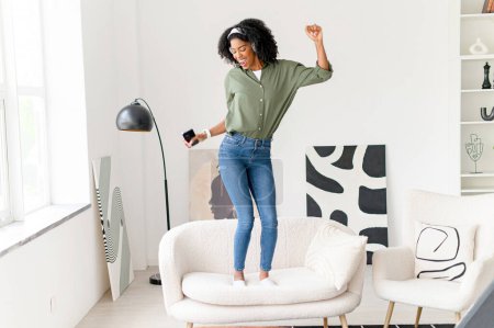 Foto de En una pose dinámica, la mujer baila en un sofá con su smartphone en la mano, auriculares puestos, totalmente inmersa en el ritmo y los latidos de su música en un espacio minimalista, moderno - Imagen libre de derechos