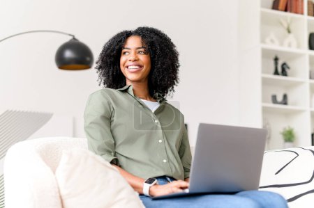 Téléchargez les photos : Une femme afro-américaine joyeuse interagit avec son ordinateur portable, son expression rayonnant de confiance et de facilité dans son espace de travail à la maison. Femme pigiste travaillant à distance - en image libre de droit