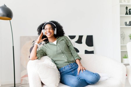 An African-American woman sits on a sofa, smiling while wearing headphones, enjoying music or a captivating audio experience. The joy of technology and the personal entertainment it provides.