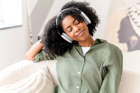 Eyes closed and with serene expression, the woman enjoys a moment of relaxation with her headphones on, embodying a peaceful break in a comfortable, modern setting. Its a snapshot of modern self-care
