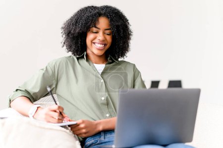 Photo for A radiant African-American woman is taking notes while working on her laptop, seated on a cozy couch in a bright, modern home setting, working or studying on the distance - Royalty Free Image