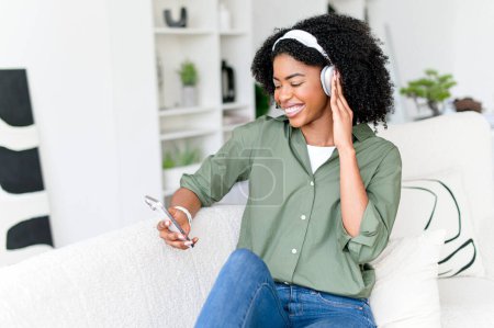 Joyful African-American woman with curly hair enjoys listening to music on her headphones and smartphone while sitting on a couch. The role of technology in contemporary relaxation and entertainment.