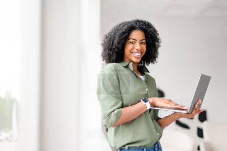 Strahlende Afroamerikanerin hantiert lässig mit Laptop, ihr fröhliches Auftreten, das den Komfort und die Bequemlichkeit des modernen digitalen Lebens widerspiegelt, unterstreicht die zeitgemäße Atmosphäre dieser häuslichen Arbeitswelt