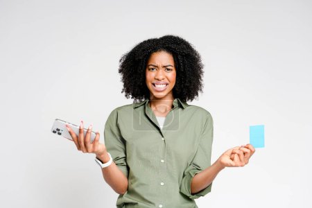Confused African-American woman holds a smartphone and a credit card, her expression of doubt perhaps indicating a problematic digital transaction or puzzling news.