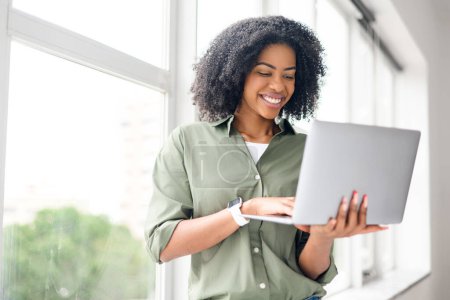 A delighted African-American woman interacts with her laptop against a backdrop of large windows, embodying the harmony and efficiency of a modern home office setting with comfort of natural lighting