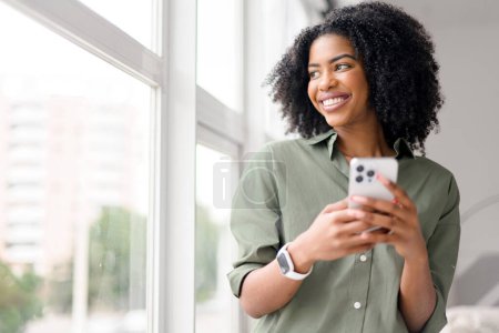 Capturando un momento de conectividad moderna, una mujer afroamericana se involucra con su teléfono inteligente, su expresión encantada enmarcada en un telón de fondo de un interior espacioso y luminoso.