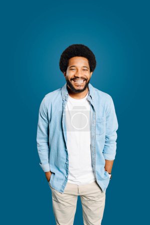 A Brazilian young man with a friendly smile and a casual denim outfit stands with a relaxed posture, hands in pockets, exuding warmth and approachability against a monochrome blue backdrop