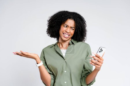 Eine afroamerikanische Frau mit verspielter Fratze blickt auf ihr Smartphone und drückt ihre Verwirrung oder Ungläubigkeit vor einem schroffen weißen Hintergrund aus, der ihre emotionale Pose unterstreicht..