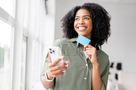 An African-American woman laughs lightly while looking at her smartphone, a blue credit card in hand suggesting a joyful online shopping experience or the ease of managing finances digitally.