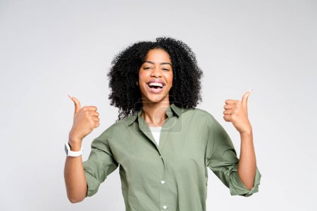 Foto de Una mujer afro-americana alegre da dos pulgares hacia arriba, exudando positividad y confianza. El retrato del estudio captura un ambiente de celebración, ideal para conceptos de éxito y felicidad - Imagen libre de derechos