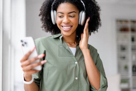Foto de Una mujer afroamericana vivaz con auriculares disfruta de una animada videollamada, su sonrisa iluminada por la luz del día que fluye a través de una ventana en un ambiente de oficina elegante y minimalista.. - Imagen libre de derechos