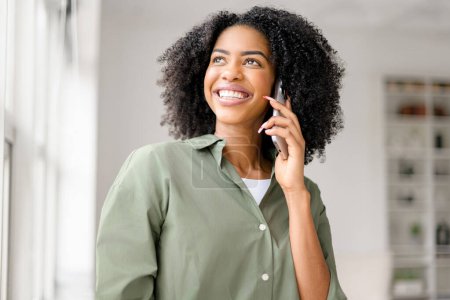 Photo for A joyful African-American woman talking on her phone, her bright smile and upward gaze suggesting good news or a delightful conversation in a cozy, sunlit space. - Royalty Free Image