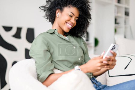 Foto de Una mujer afroamericana encantada se relaja con su teléfono, desplazándose por las redes sociales, con el telón de fondo de una decoración hogareña elegante y minimalista. La tecnología se mezcla a la perfección en la vida cotidiana - Imagen libre de derechos