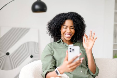 Téléchargez les photos : Avec une vague ludique et un sourire captivant, la femme afro-américaine semble accueillir quelqu'un lors d'un appel vidéo sur son smartphone, entouré par le confort de sa maison moderne et minimaliste - en image libre de droit