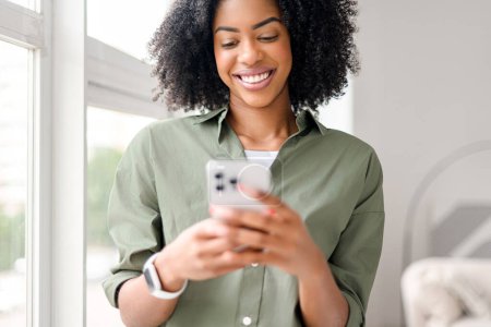 Sourire engageant et smartphone à la main, la femme afro-américaine semble profiter d'une conversation chaleureuse ou recevoir de bonnes nouvelles, le tout dans le confort d'un environnement familial contemporain