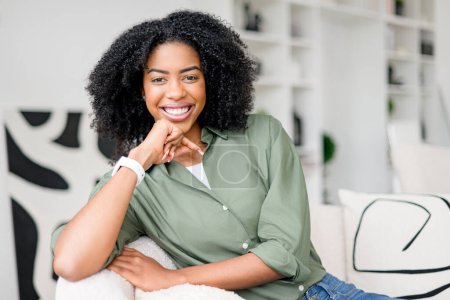 Smiling broadly, an African-American woman sits on a fluffy white sofa, her hand gently touching her face, in a scene that conveys casual elegance and a relaxed, comfortable home environment.