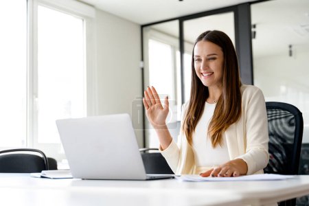 A young, cheerful businesswoman greets her virtual audience with a wave during an online meeting, her radiant smile and the bright office setting reflecting a welcoming and positive corporate culture