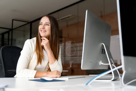Captured with a grin, a young female office employee enjoys her work on a desktop computer, reflecting the comfortable and employee-friendly atmosphere of a modern workplace. Job satisfaction concept